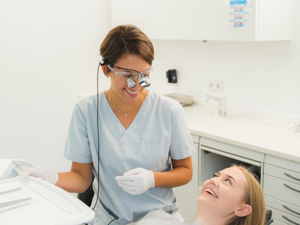 Zahnärztin mit Lupenbrille steht lächelnd neben einer Patientin im Behandlungsstuhl in einem hellen Behandlungsraum